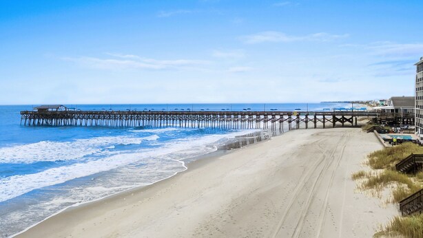 A beach with a wooden pier extending into the ocean, a sandy beach, and a building with a balcony overlooking the beach. - Inlet Paradise B Beautiful Condo Walking Distance to Beach! (Murrells Inlet)