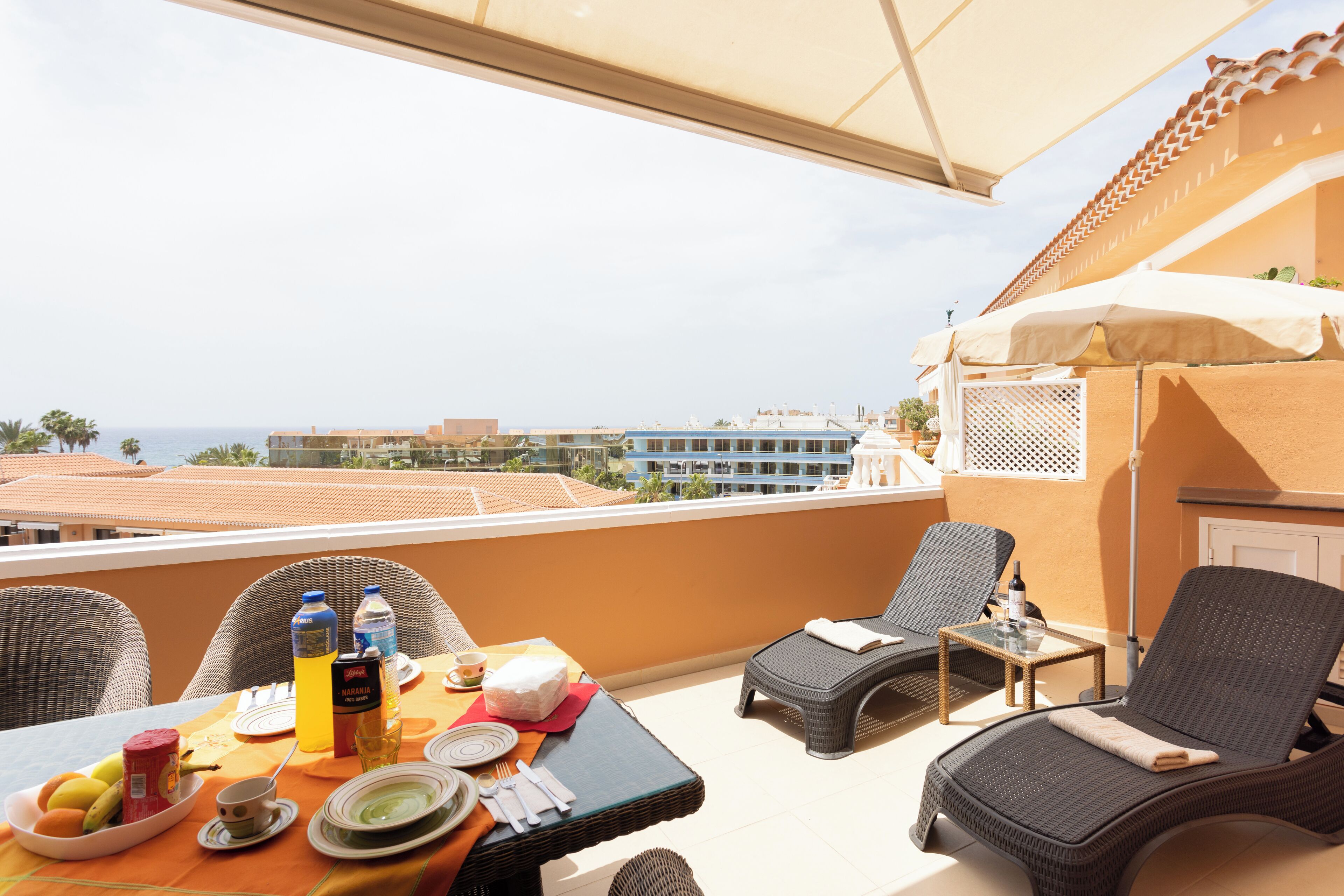 A balcony with wicker chairs, a table set for breakfast, and a view of the sea and buildings.
