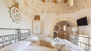 Een slaapkamer met een stenen plafond, een bed met witte lakens, een aan de muur bevestigde televisie en een boogvormige deuropening naar een andere kamer.
