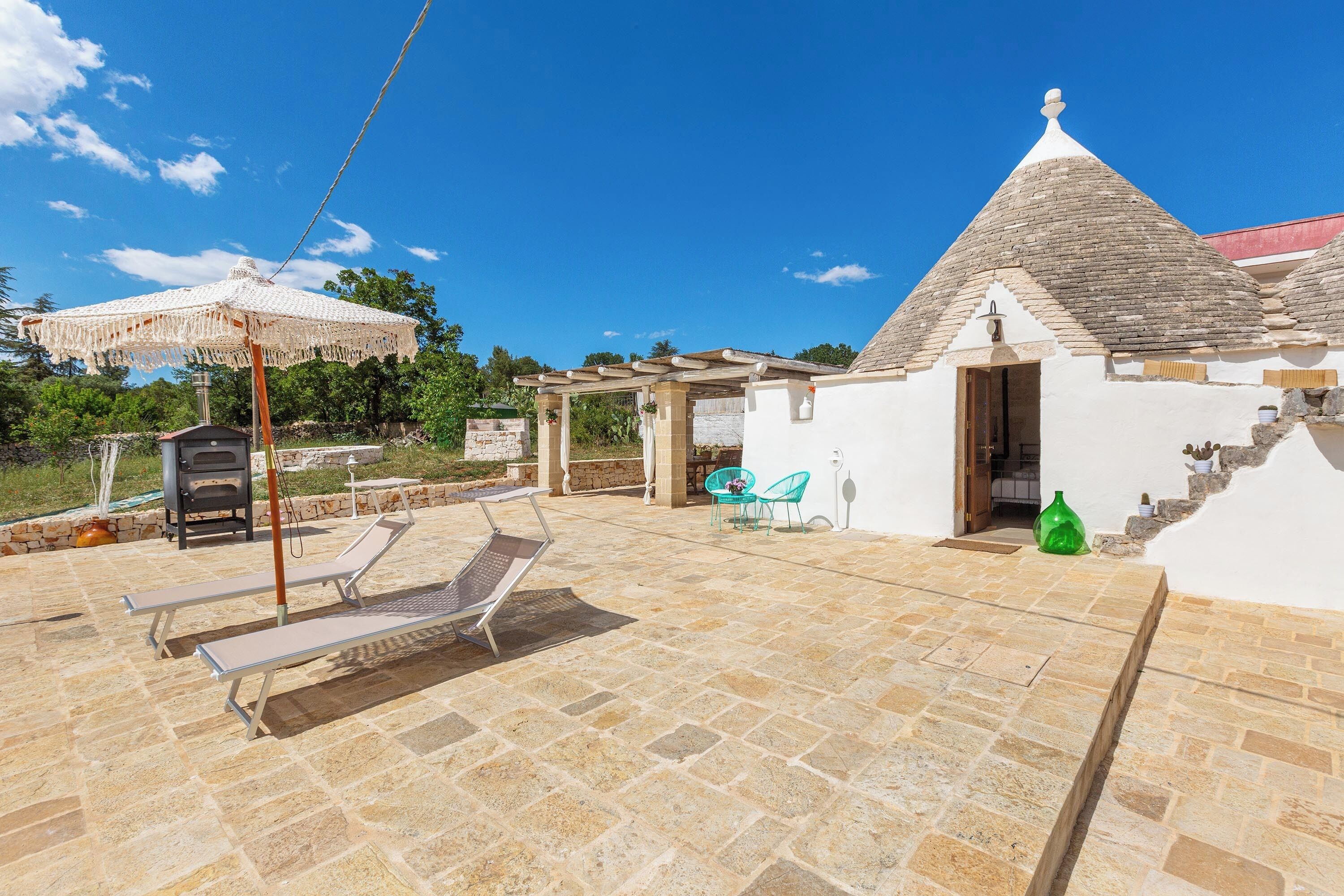 A patio area with stone flooring, lounge chairs, a pergola, and a small building with a tiled roof.
