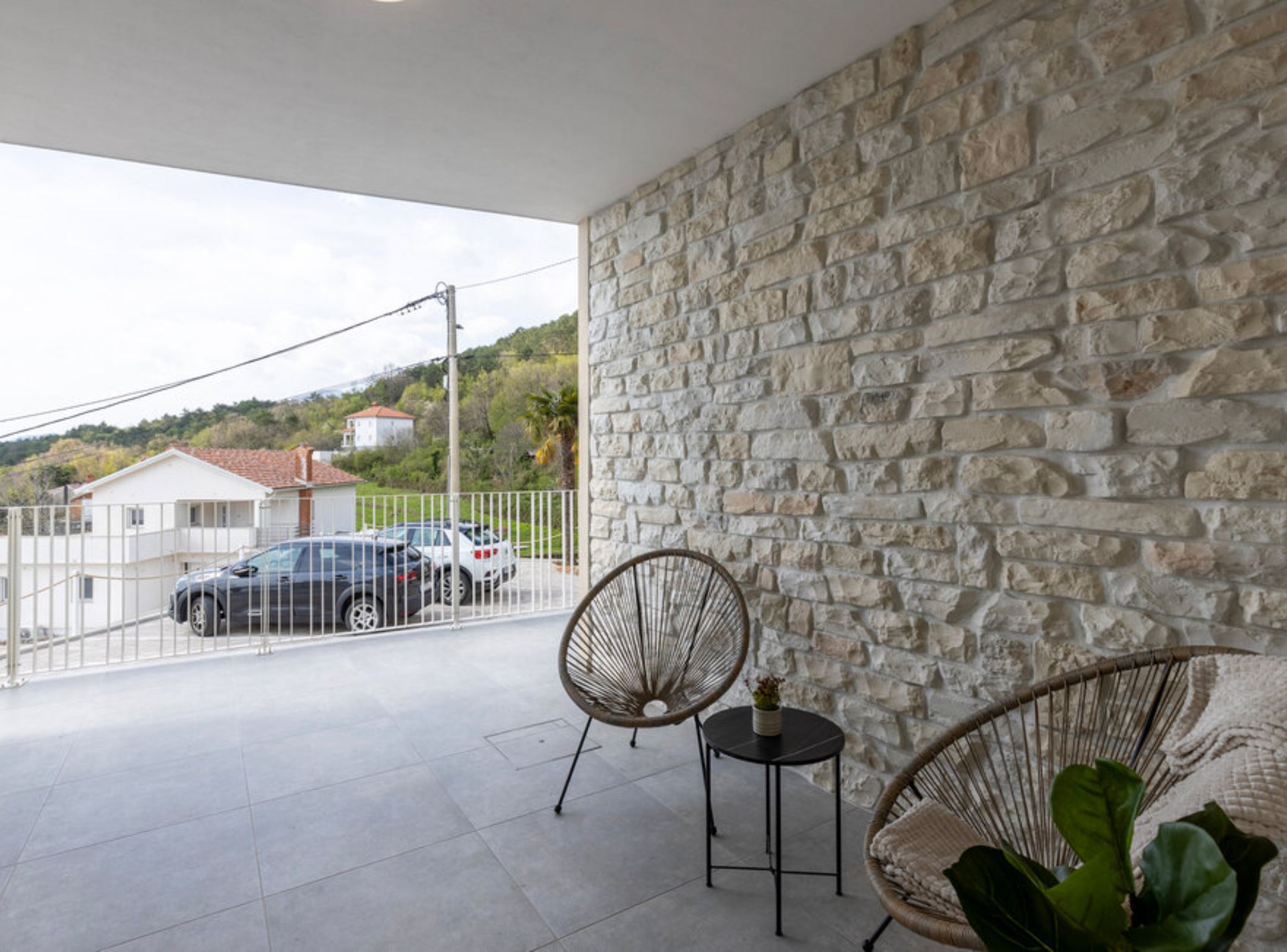A balcony with stone walls, two wicker chairs, a small table, and parked cars in the background.