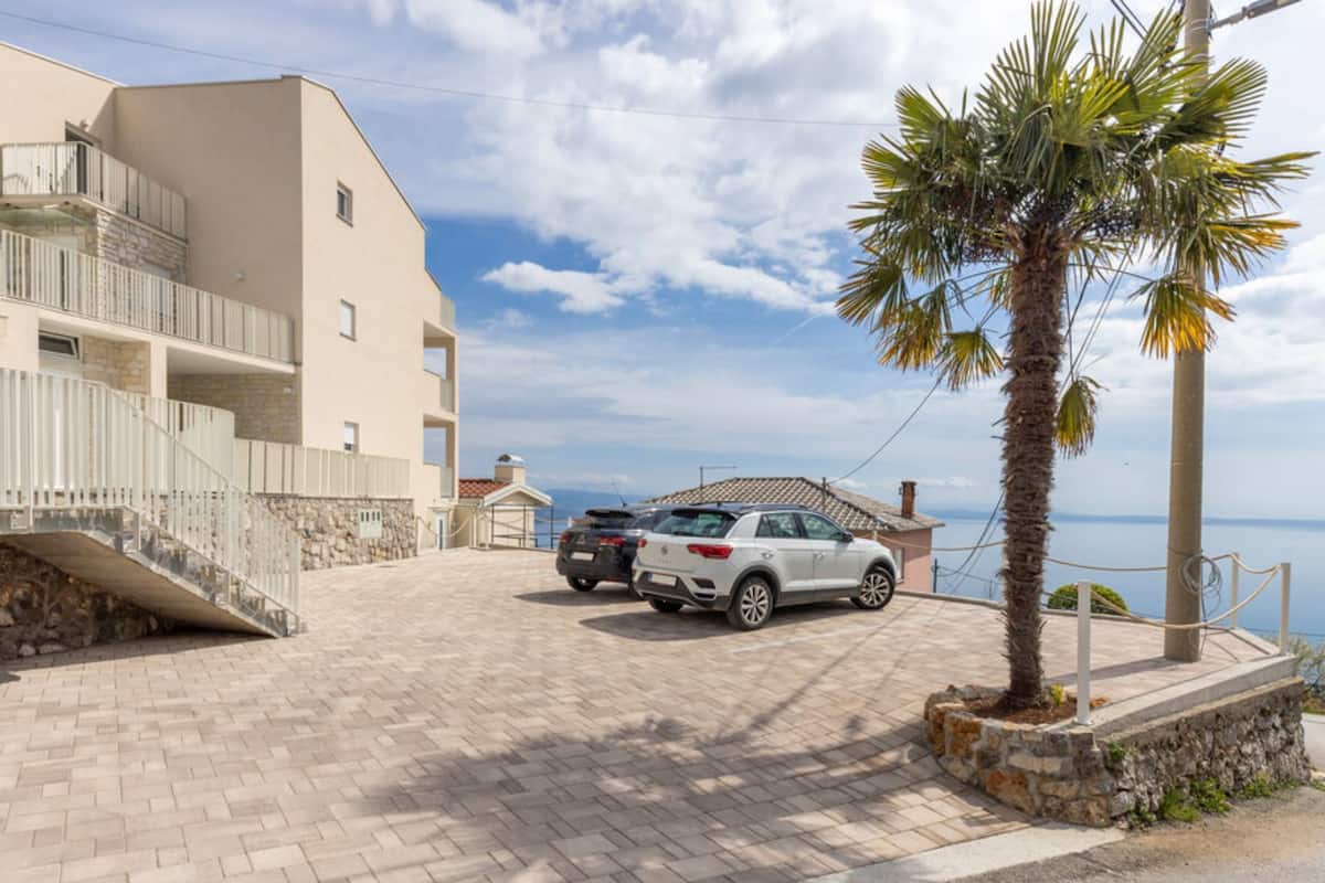 A residential building with a paved driveway, a white SUV parked in front, and a palm tree to the right.
