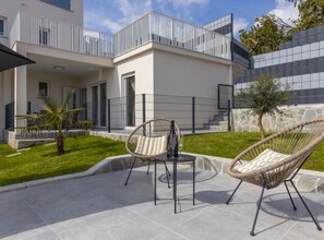 A modern outdoor patio with two wicker chairs and a small table, a white building with balconies, and a stone wall. - Apartments Bregha (Matulji)