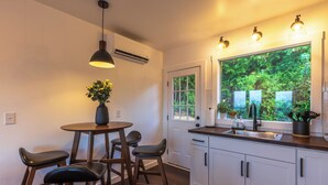 A modern kitchen with a round wooden table, bar stools, a large window with a view of greenery, and white cabinets.