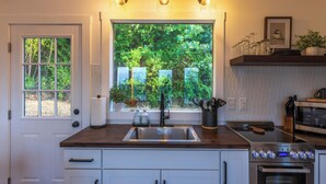 A kitchen with a large window, a stainless steel sink, a stove, and overhead lighting.