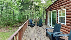 Eine Holzterrasse mit zwei Adirondack-Stühlen, eine Glastür, die zu einem Raum führt, und ein bewaldeter Hintergrund.