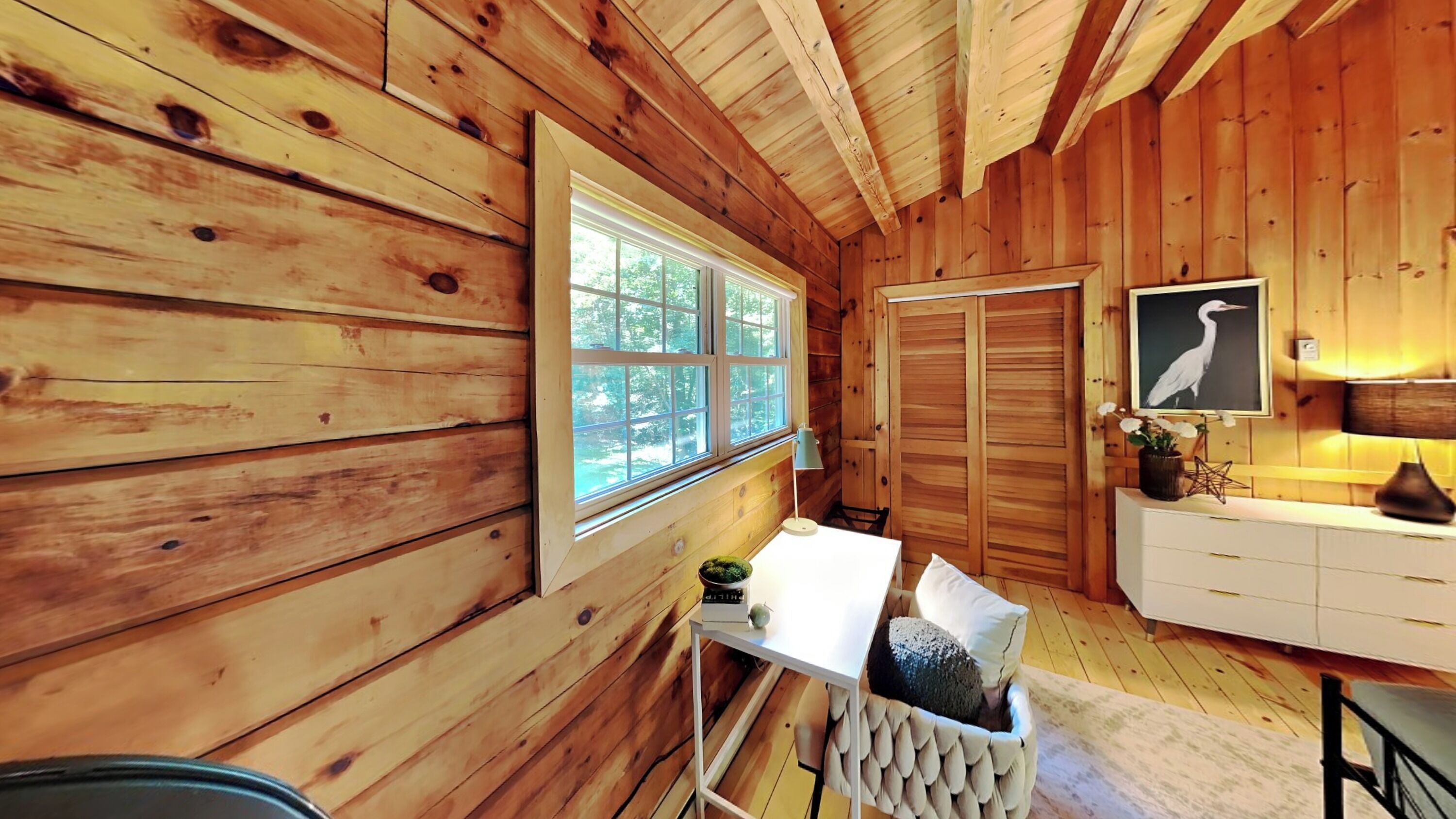 Interior de una cabaña de madera con comedor, una mesa blanca, un banco, un radiador y un ventanal grande que proporciona luz natural.