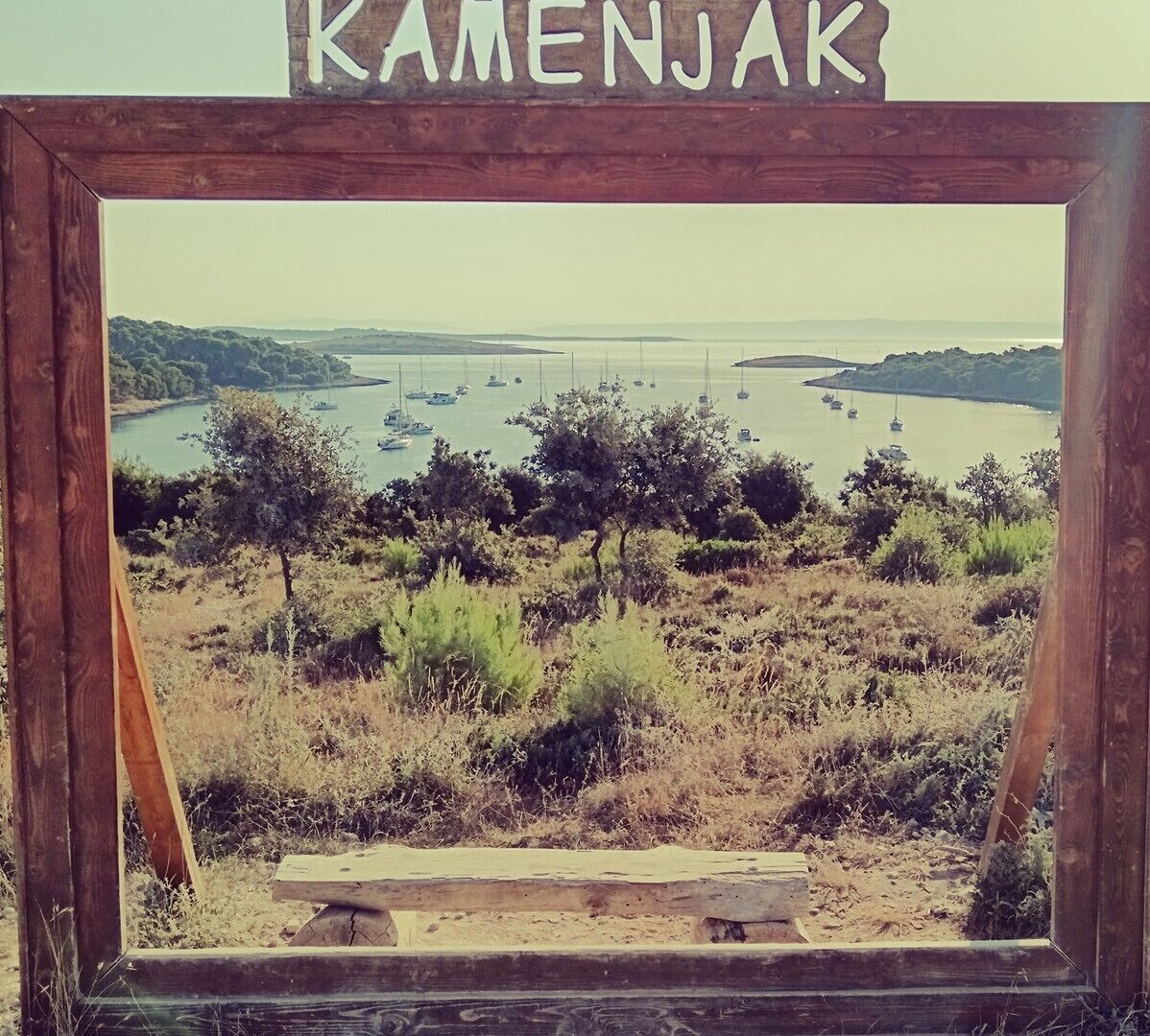 Ein Rahmen zeigt eine Küstenlandschaft mit Booten auf dem Wasser, Bäumen und einer hölzernen Bank.
