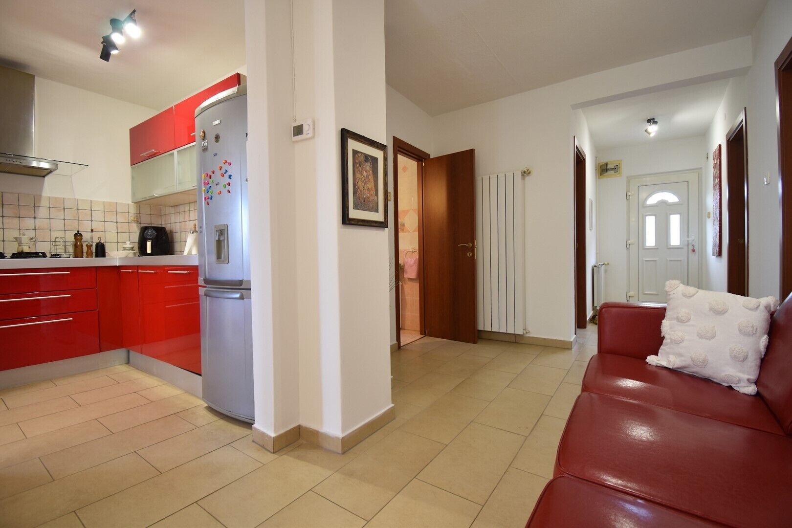 A modern kitchen with red cabinets, a stainless steel refrigerator, and a tiled floor. There is a red leather sofa with a decorative pillow in the living area.