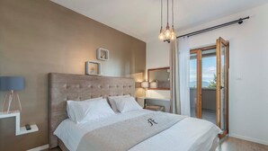 A bedroom with a large bed, a bedside table with a lamp, a nightstand, and a view of the outdoors through a sliding door.