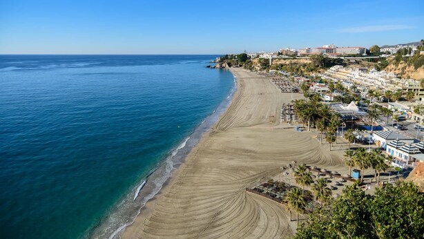A coastal town with a wide sandy beach, clear blue sea, and buildings along the shore. - Paco Romo (Nerja)