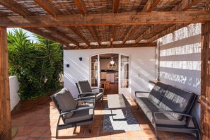 An outdoor patio with a wooden pergola, a sofa, two chairs, a table, and a kitchen area visible through an archway. - Paco Romo (Nerja)