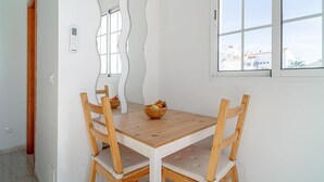 A dining area with a wooden table and chairs, a bowl of fruit, a wall-mounted thermostat, and a window with a view of buildings. - Edf Papillon (Nerja)