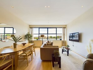 A modern living room with a sofa, dining table, chairs, a TV mounted on the wall, and large windows.