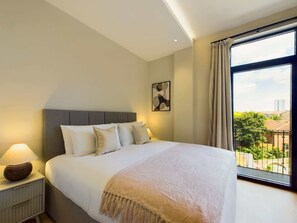 A bedroom with a large bed, a bedside table with a lamp, a window with curtains, and a city view.