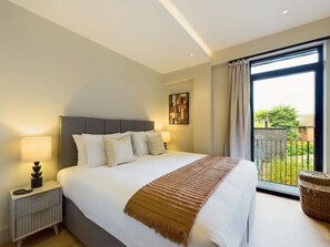 Un dormitorio con cama, lámparas de noche, una obra de arte en la pared, un balcón con vistas y una cesta de mimbre.