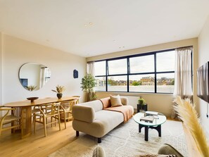Ein modernes Wohnzimmer mit Sofa, Couchtisch, Esstisch, Stühlen, einem großen Fenster und einem Flachbildfernseher.