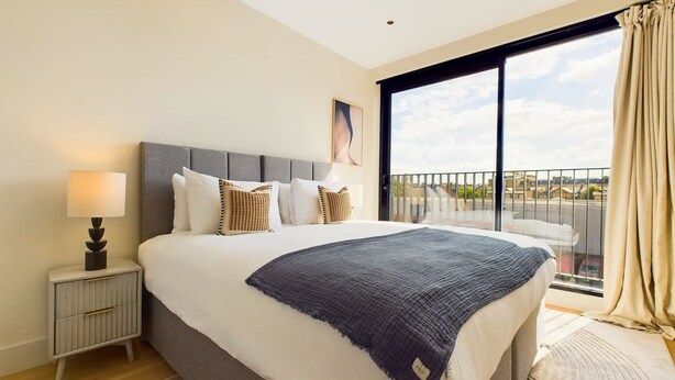 A hotel room with a large bed, a bedside table with a lamp, a view of the city through a balcony. - Warm Relaxing St Pancras Apartment with Balcony (Greater London)