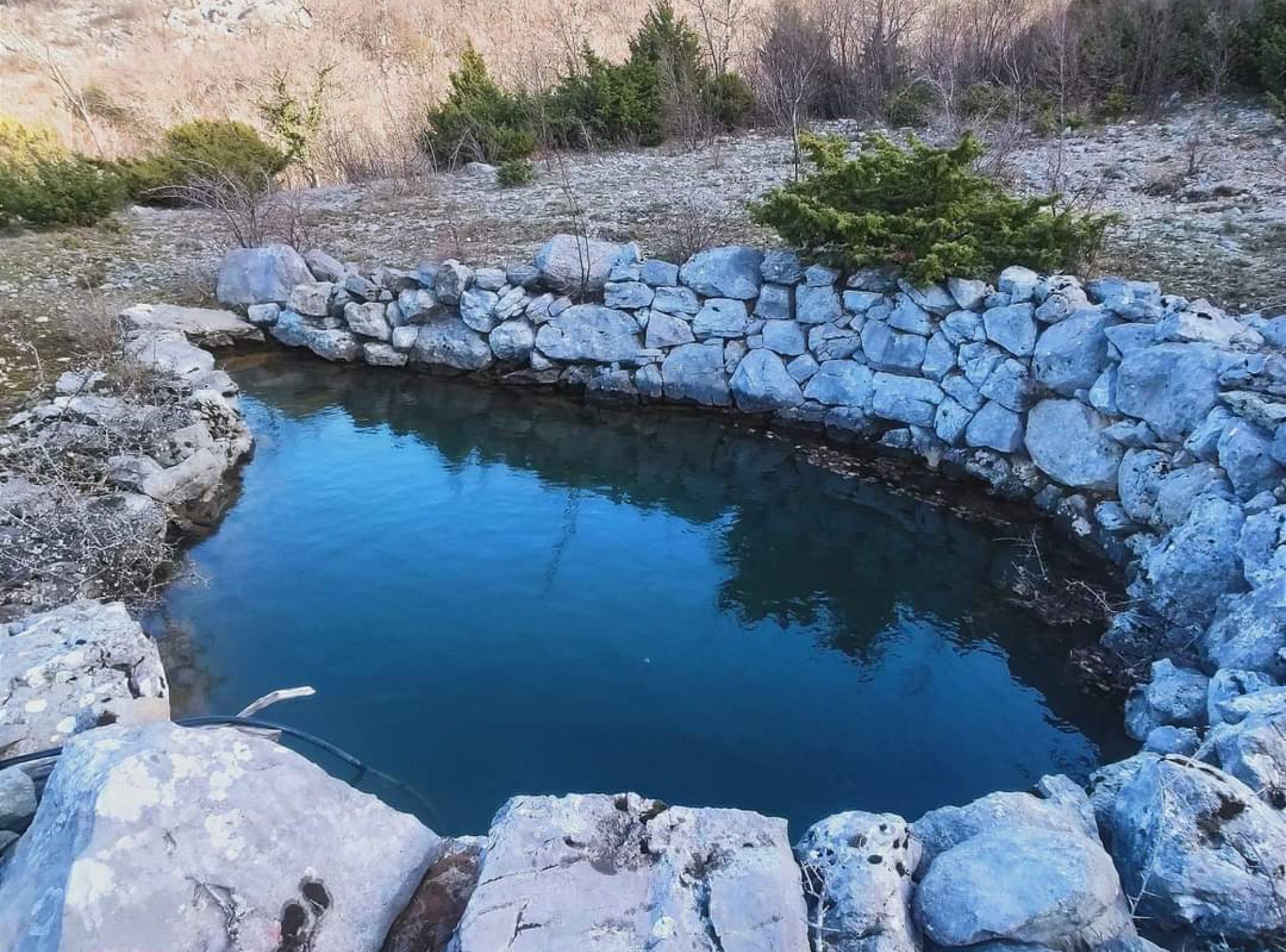 Un petit étang aux eaux claires, entouré d’un mur de pierres, avec une végétation clairsemée et un paysage aride et rocheux en arrière-plan.