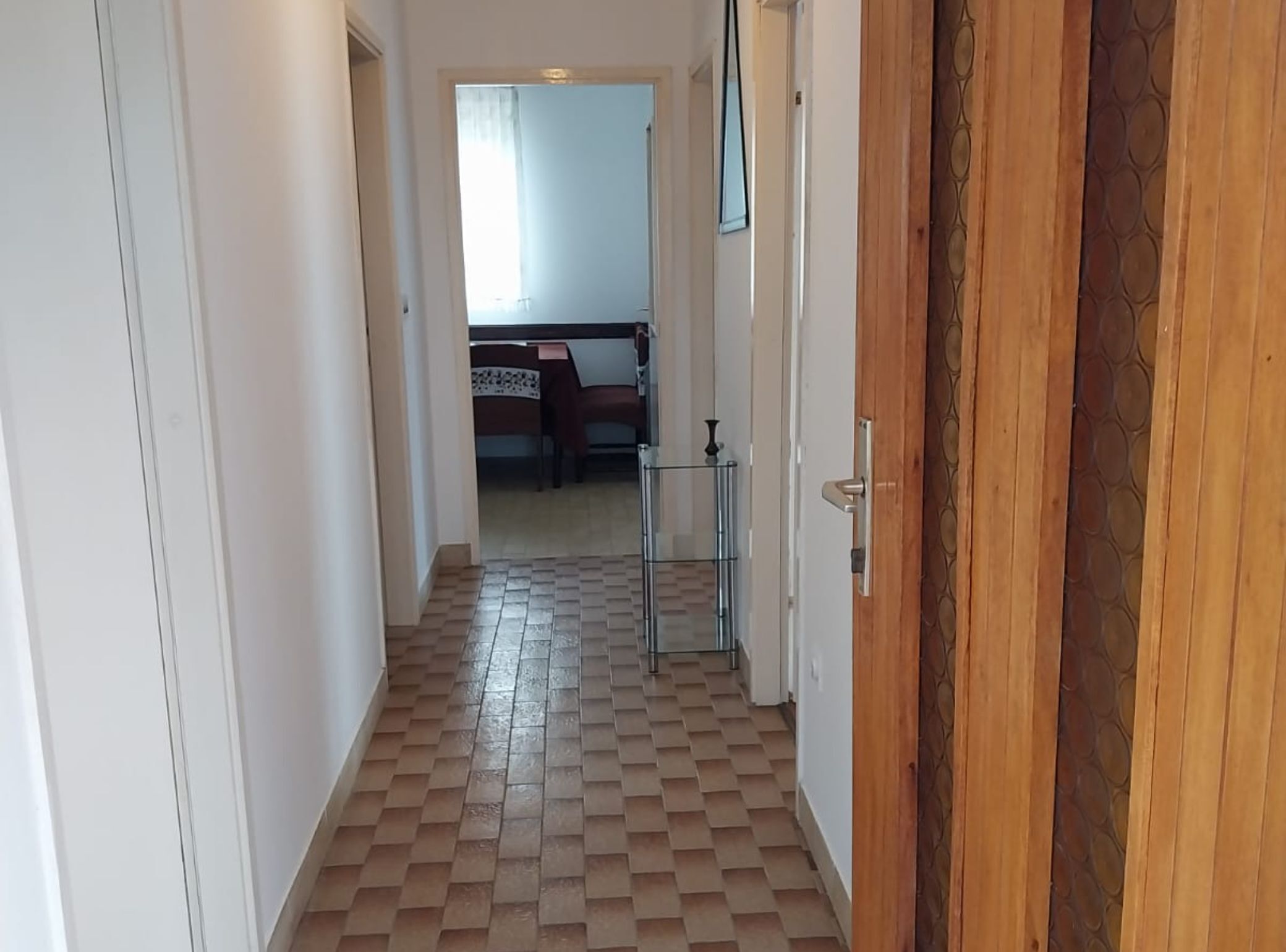 A hallway with a checkered floor leading to a room with a bed and a chair.