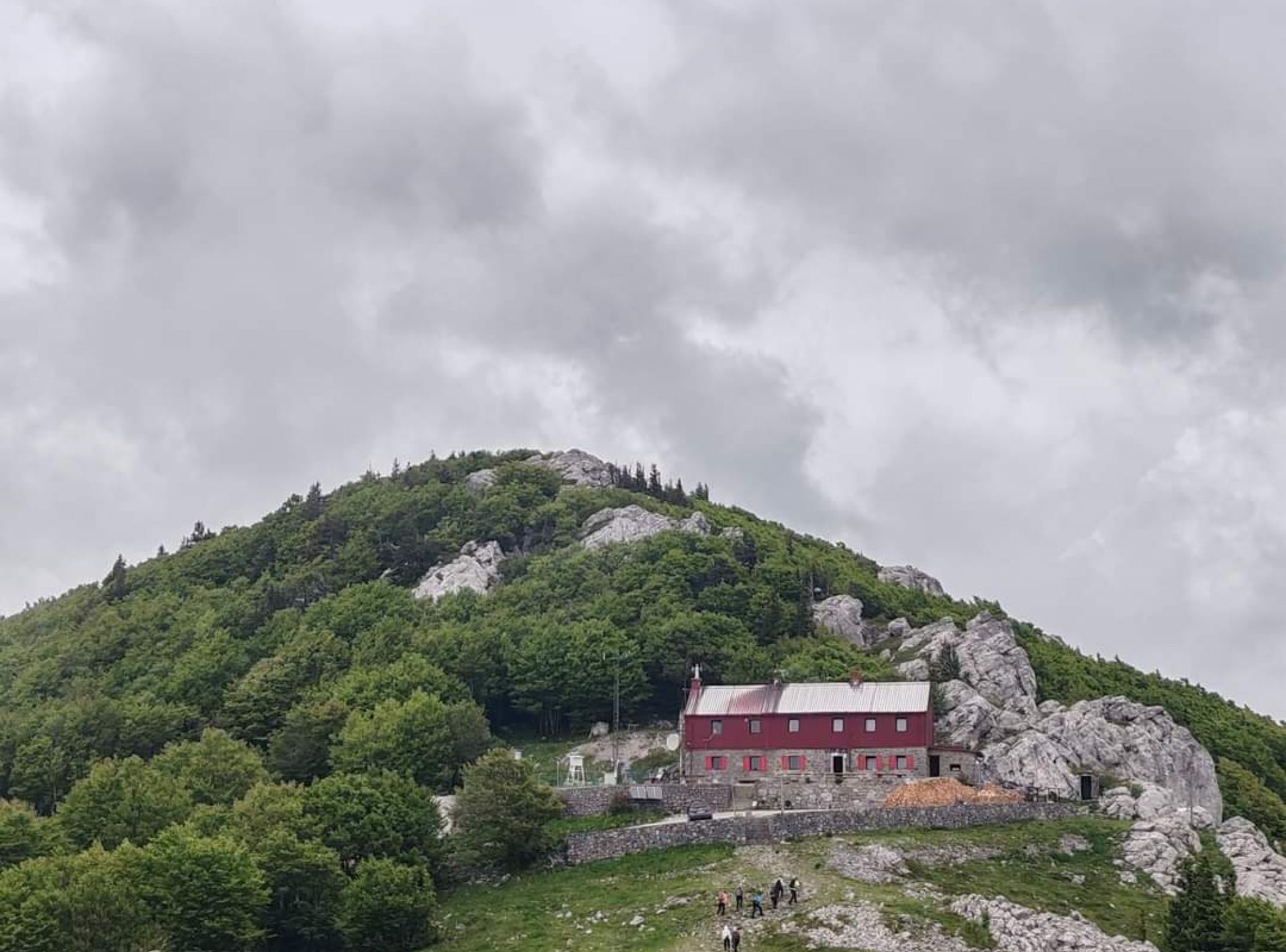A red building with a stone foundation on a hillside, surrounded by greenery and rocks.