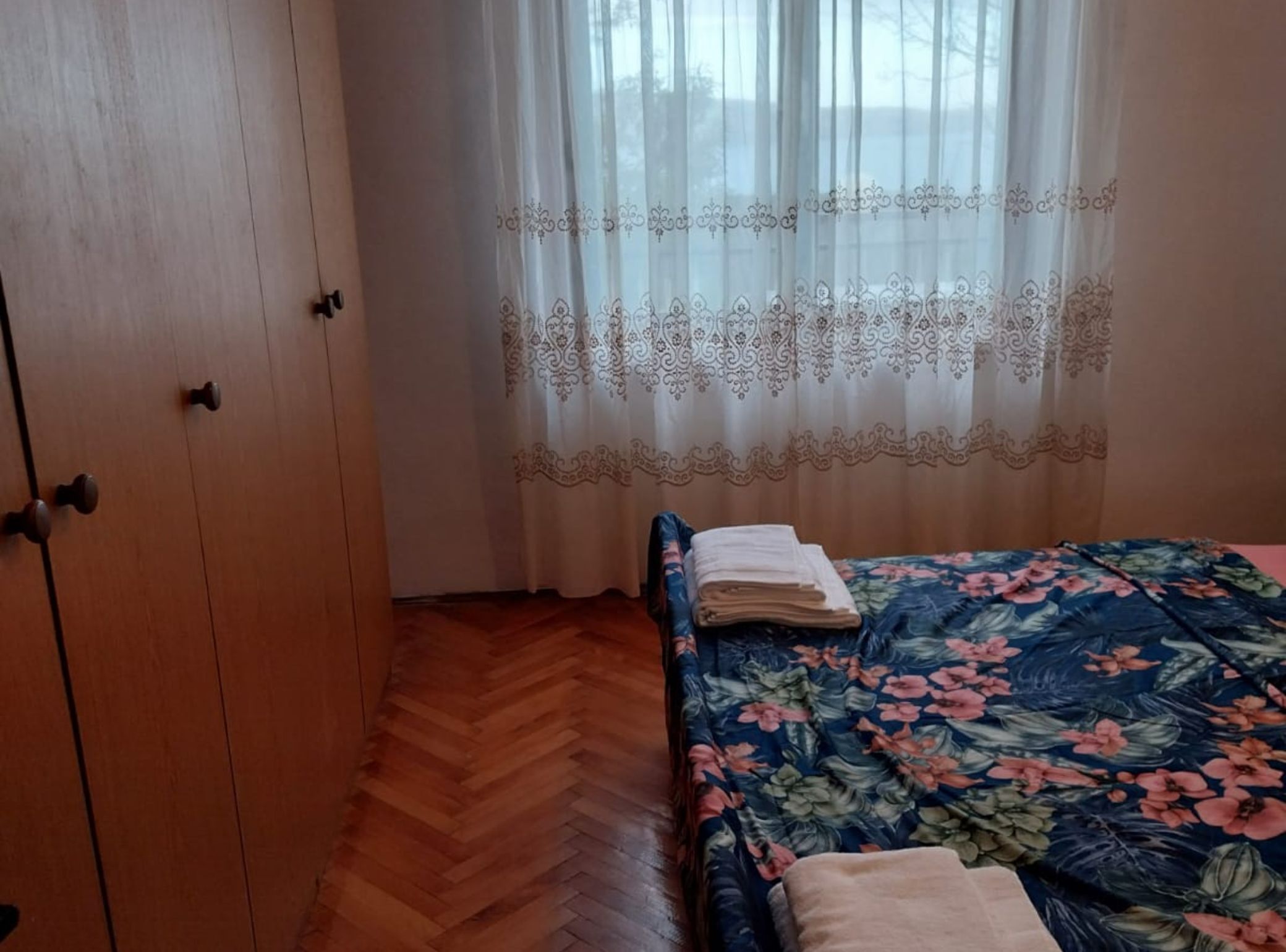 Un dormitorio con un armario de madera, una cama con colcha estampada y una ventana con cortinas traslúcidas.