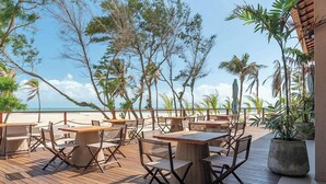 An outdoor deck with wooden tables and chairs, surrounded by tropical plants and overlooking a beach. - Vila Galé Collection Sunset Cumbuco (Caucaia)