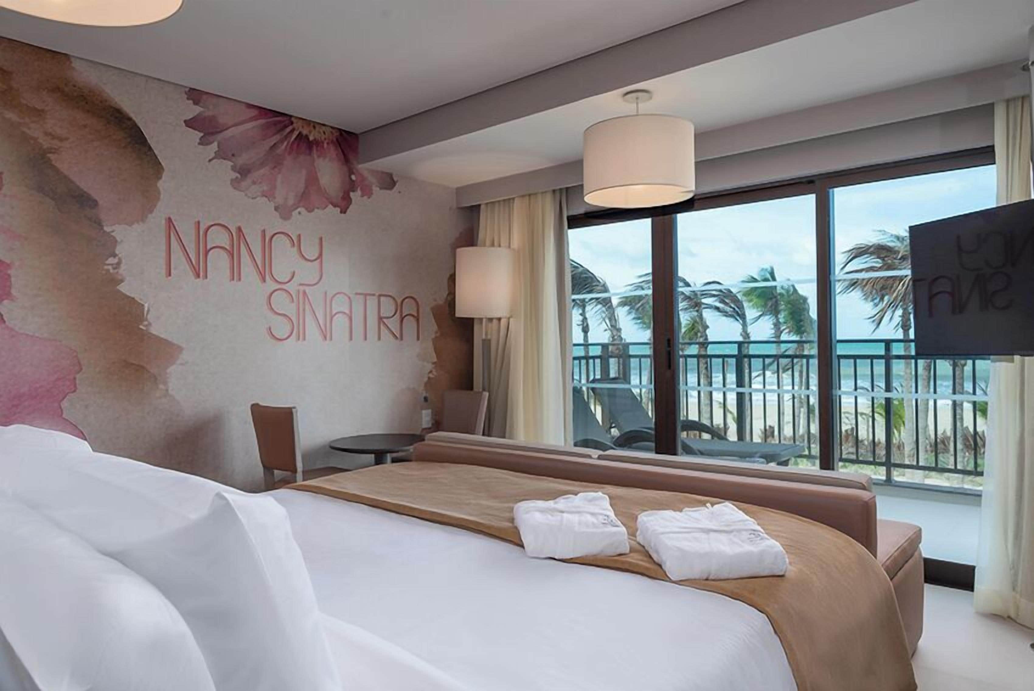 A hotel room with a bed, a desk, a chair, a TV, and a balcony with a view of palm trees and the ocean.