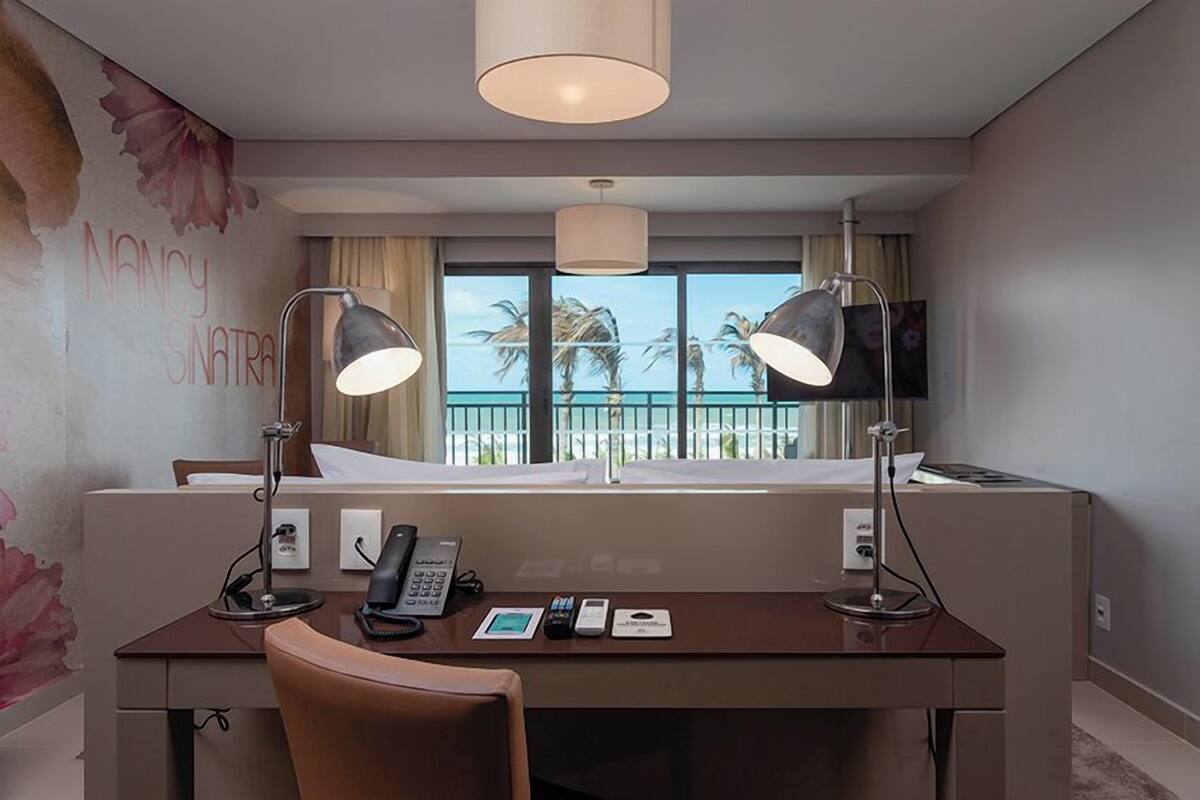 A hotel room with a desk, two lamps, a phone, a small table, and a window with a view of palm trees and the sea.