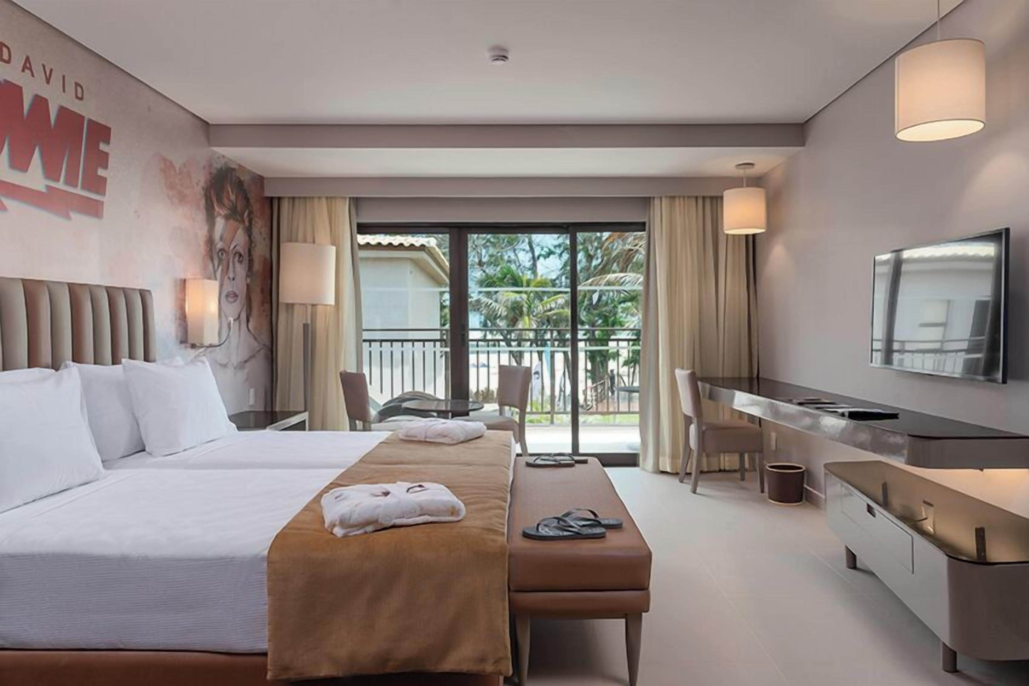 Quarto de hotel com duas camas, uma escrivaninha, um banco, uma televisão e varanda com vista para palmeiras.