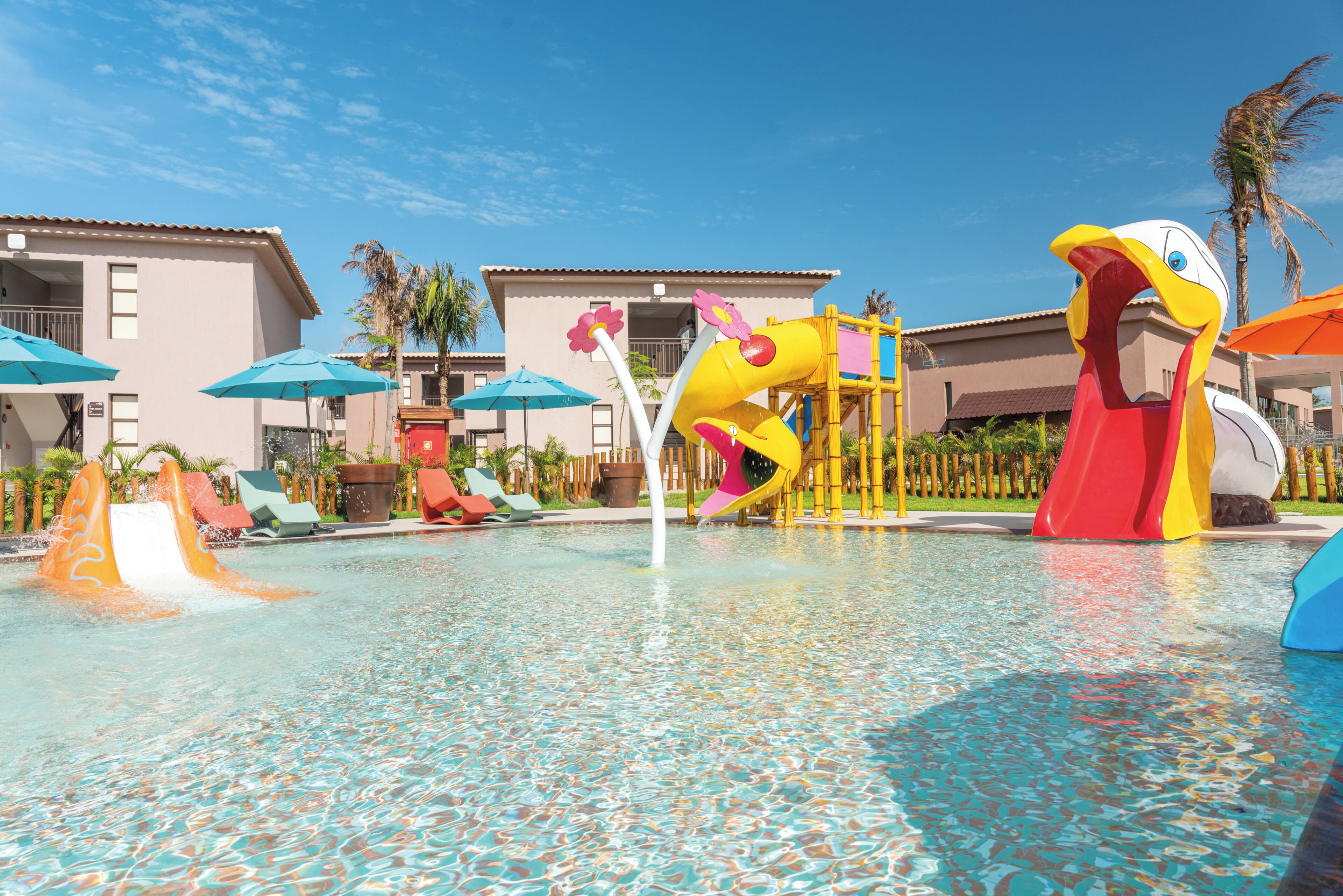 A swimming pool with slides and a water play area, surrounded by buildings with balconies and palm trees.
