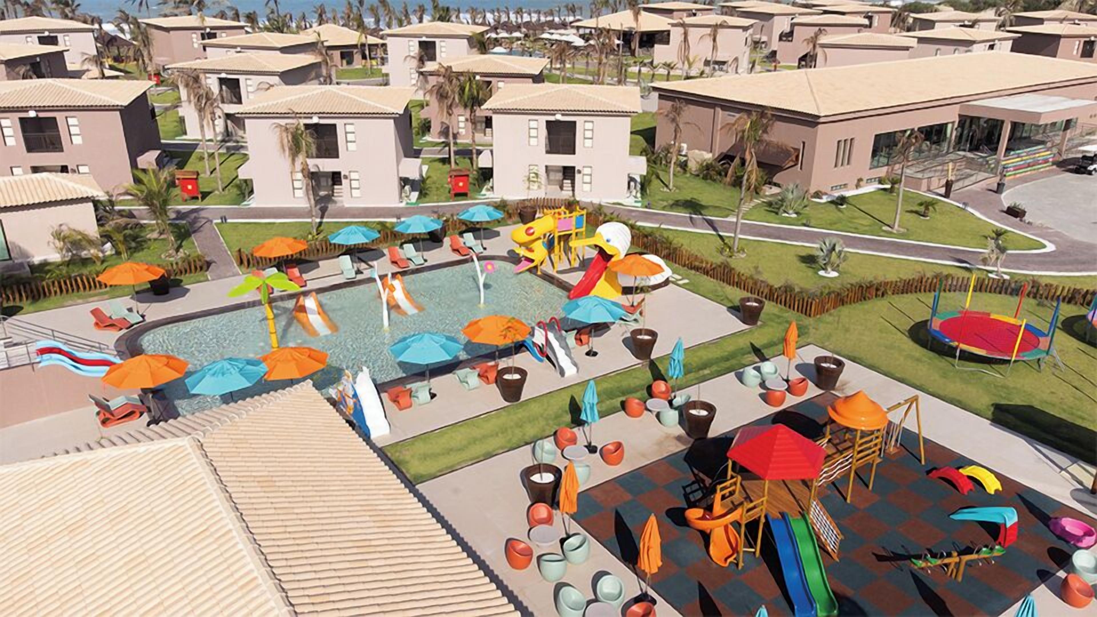 Un complejo turístico con varias edificaciones, un área de piscina central con toboganes y sombrillas, y un parque infantil con toboganes y columpios.