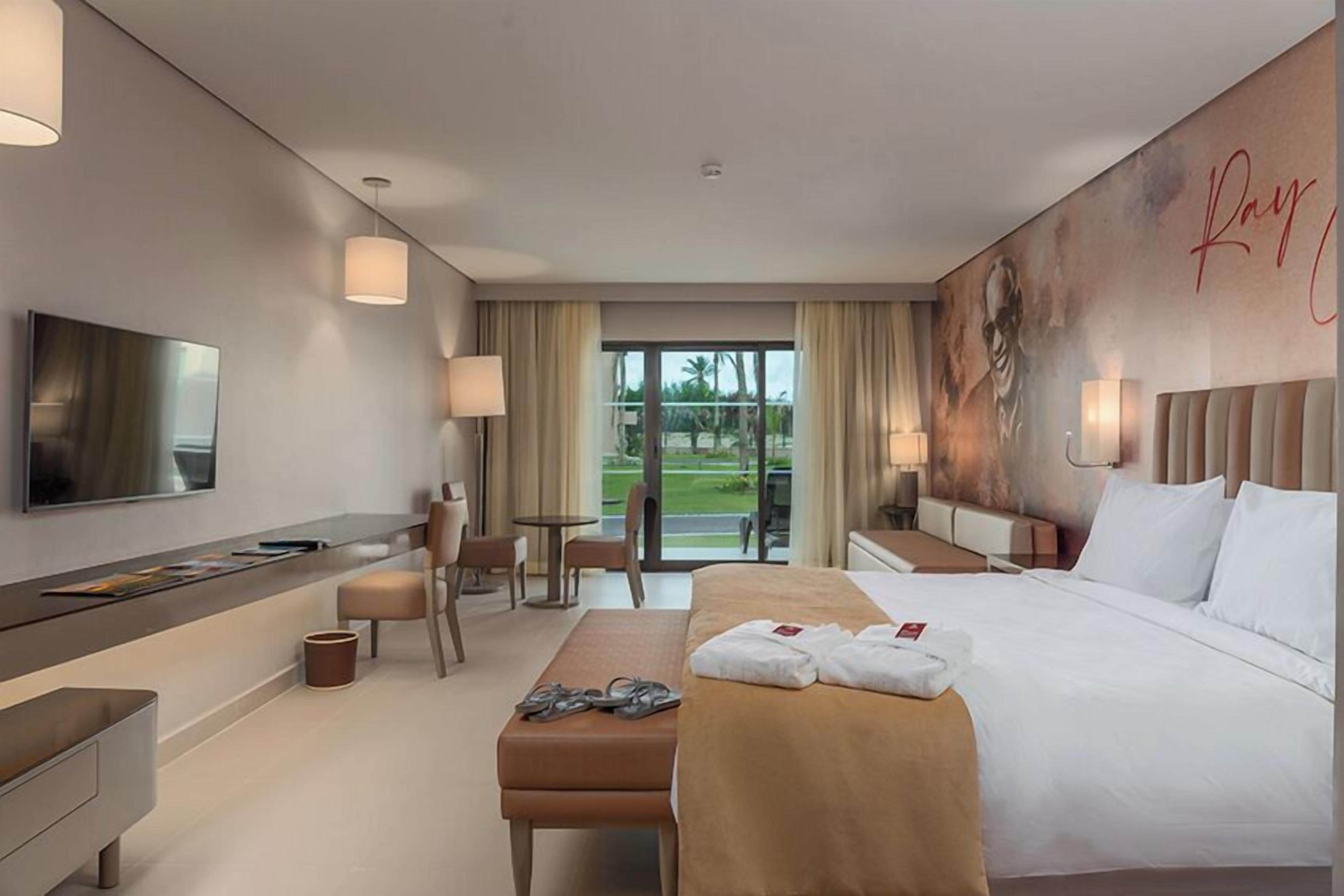 A modern hotel room with a large bed, a desk, a TV, and a view of the outdoors.