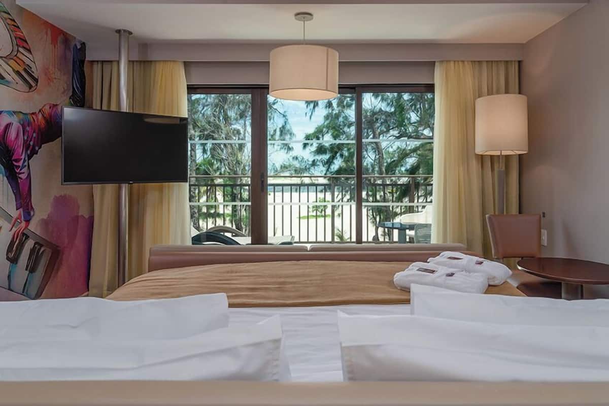 A hotel room with a bed, a TV, a balcony with a view of palm trees, and a painting on the wall.