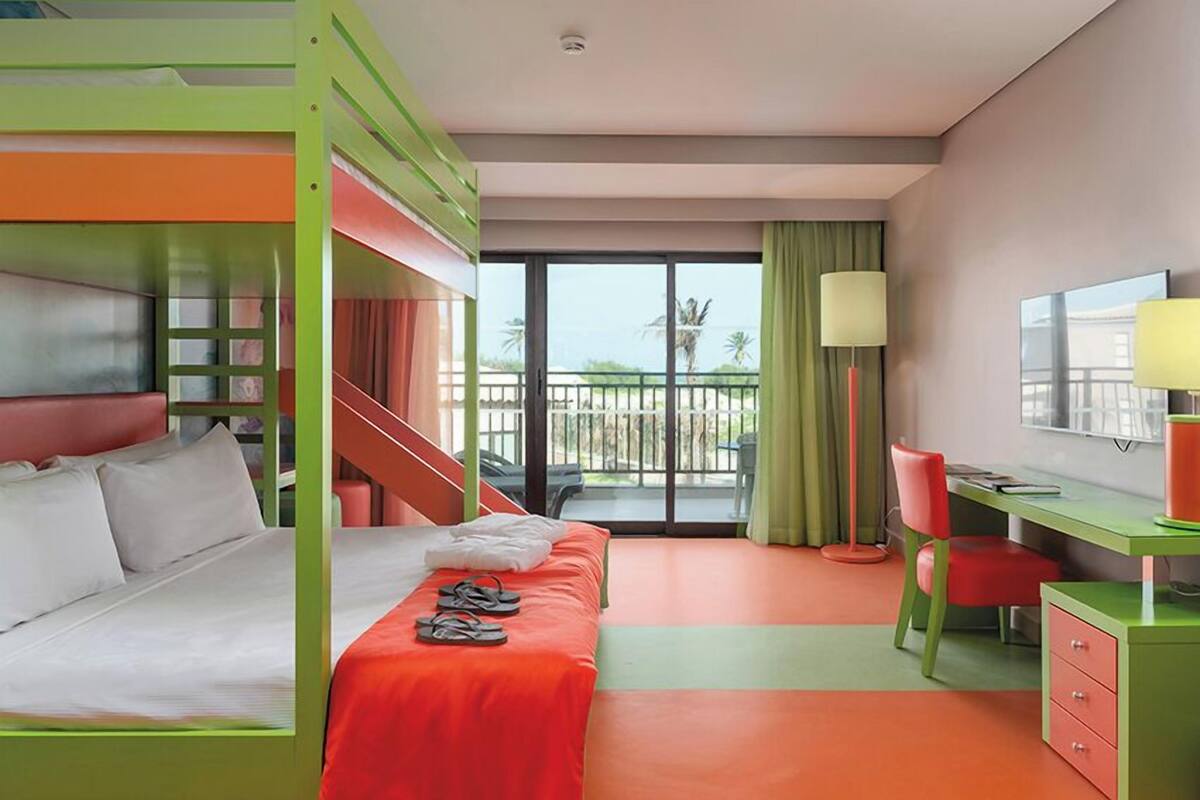 A hotel room with a bunk bed, a desk, a chair, a lamp, and a view of the balcony.