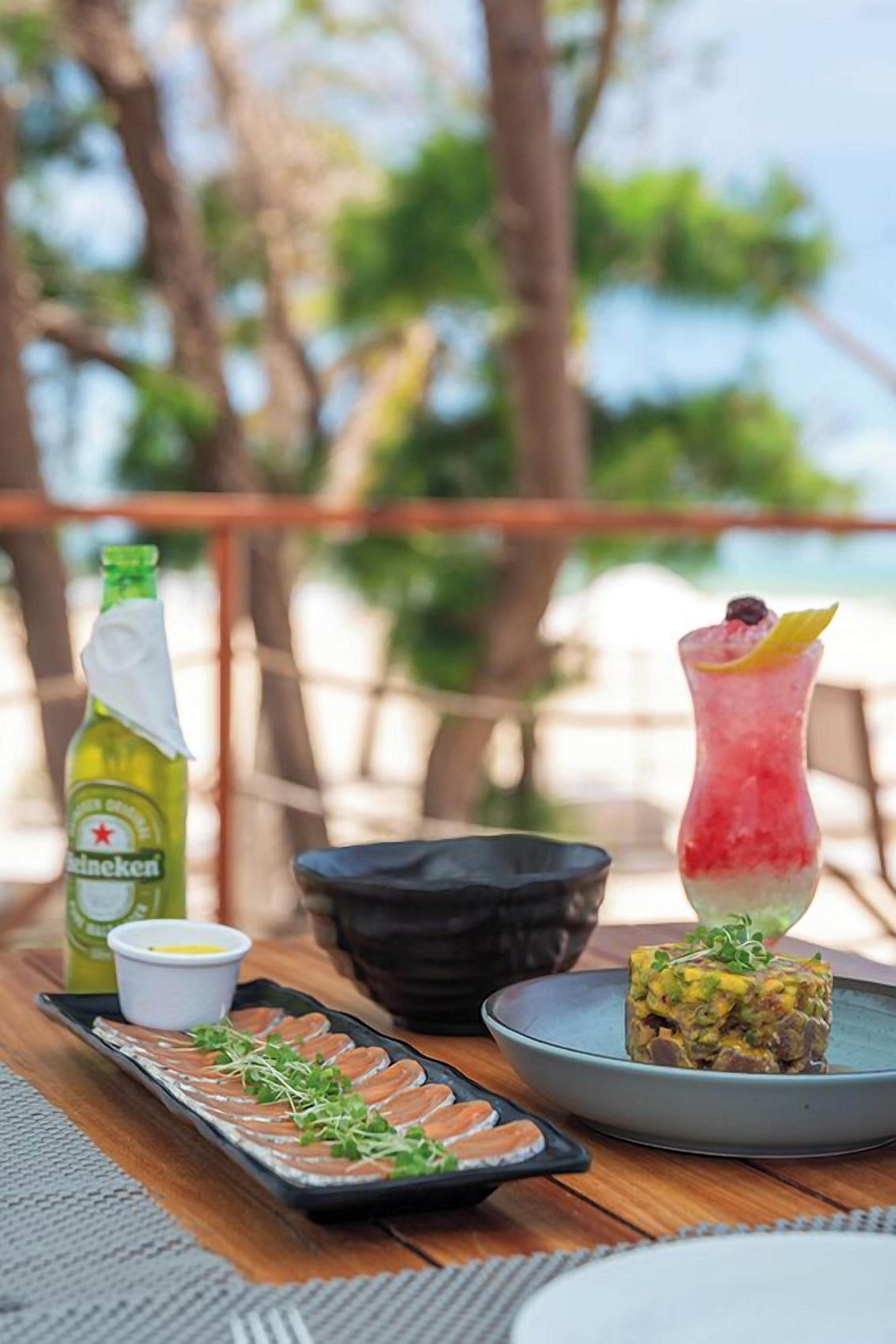 Une table à manger avec une bouteille de Heineken, une assiette de sashimi, un grand verre rempli d’une boisson décorée d’une tranche de citron et un petit bol de sauce.
