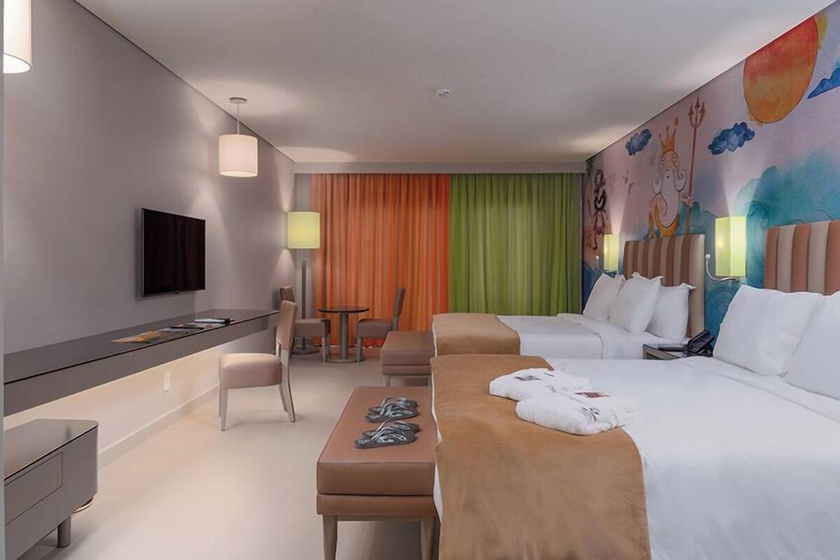 A hotel room with a flat-screen TV, a desk with a chair, a massage table, and a bed with pillows and a towel.
