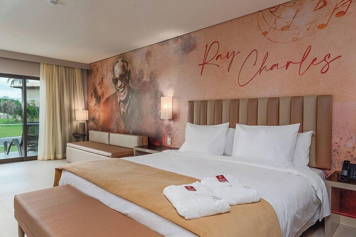 A hotel room with a large bed, a bench, a desk with a lamp, and a view of the outdoors.