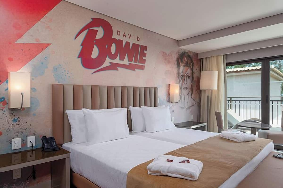 A hotel room with a large bed, a headboard, a bedside table with a phone, a lamp, and a wall with a David Bowie mural.