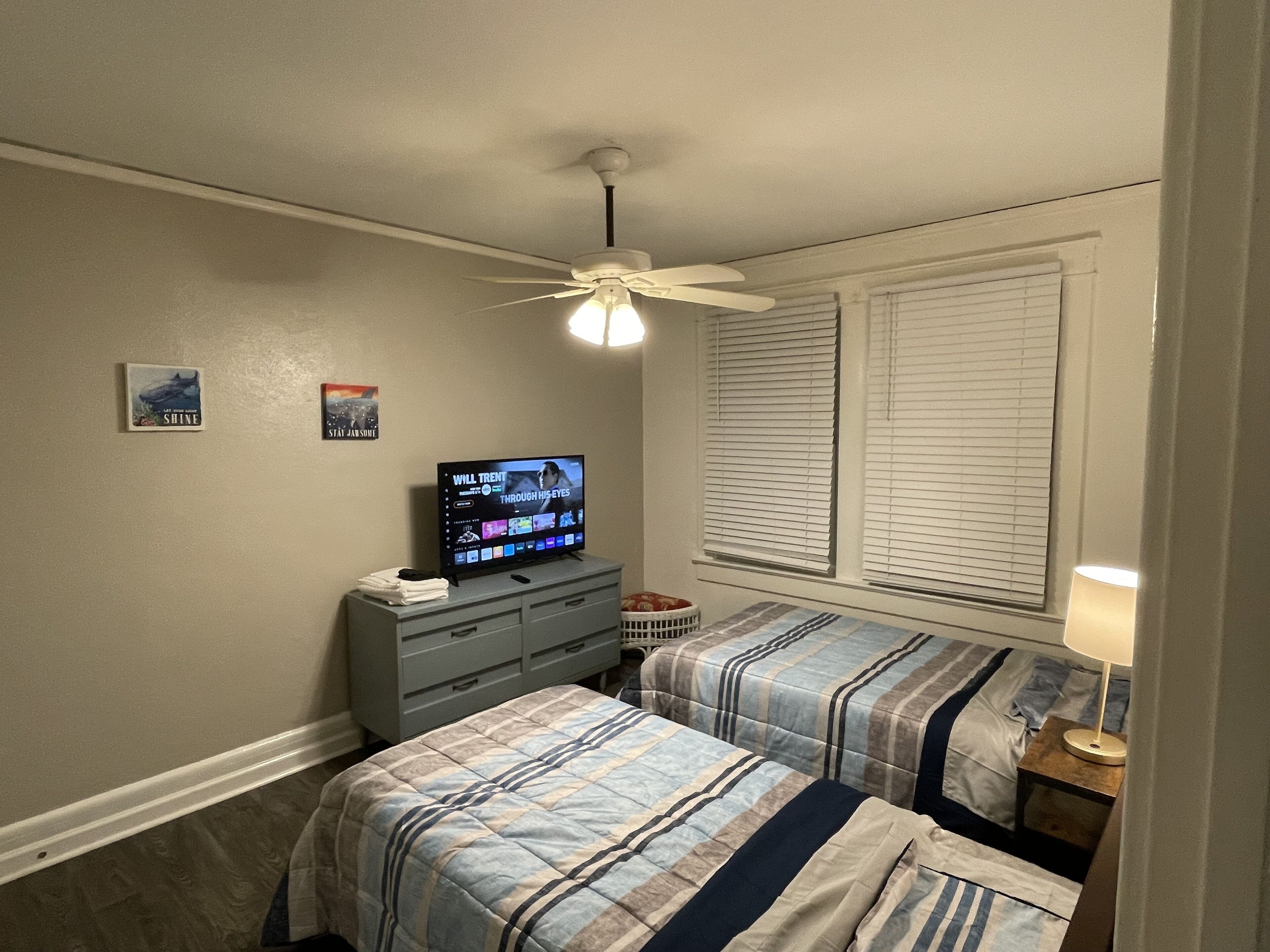 Une chambre avec deux lits, un ventilateur de plafond, un téléviseur posé sur une commode et une fenêtre avec des stores.