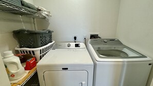 A laundry area with a front-loading washer and dryer, a basket, and a shelf with laundry supplies. - Yellow Door Retreat I Mountain Views I Privacy I 30 Minutes to Moab! (La Sal)