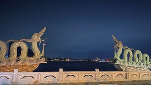 A night view of a bridge with dragon sculptures on either side, overlooking a body of water with city lights in the distance.