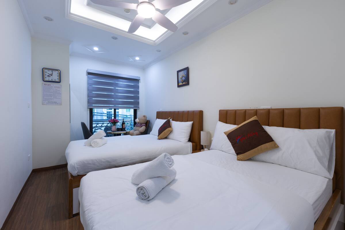 A hotel room with two beds, a ceiling fan, a window with blinds, and a clock on the wall.