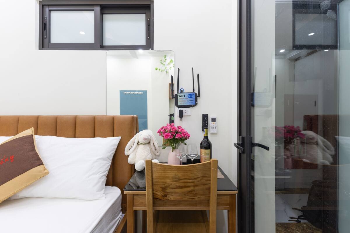 A hotel room with a bed, a wooden chair, a small table with a bottle, glasses, and a stuffed toy.