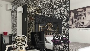 Ein Schlafzimmer mit Wand in Schwarz-Weiß-Muster, einem Bett mit Kopfteil, einem Schreibtisch mit Computer, einem Stuhl und einer kleinen Ablage mit einer roten Schale.