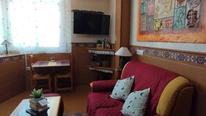Una sala de estar con un sofá rojo, un televisor montado en la pared, una mesita con una lámpara y un cuadro decorativo sobre el sofá.