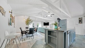 A spacious living area with a high ceiling, a central island with a sink, a dining area with chairs, and a TV mounted on the wall.