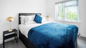Une chambre à coucher comprenant un lit, deux tables de chevet, une fenêtre avec des stores et une vue sur la verdure à l’extérieur.