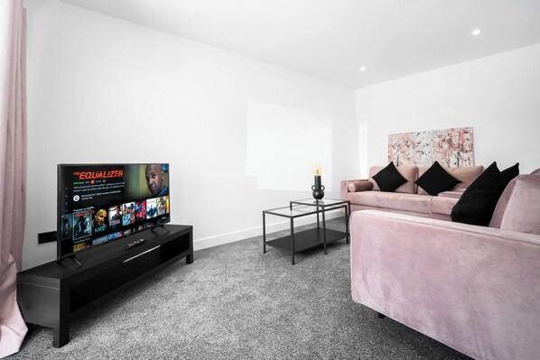 Una sala moderna con un televisor de pantalla plana, una mesa de centro de vidrio, un sofá rosa y un cuadro floral en la pared.