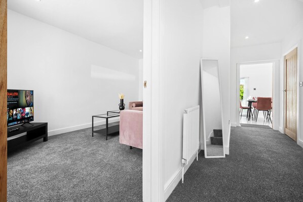 A modern hotel room with a grey carpet, a TV on a black stand, a pink chair, a small table, and a white door leading to a dining area. - Family/contractors 3 bed House/parking/sleeps 7 (Bilston)